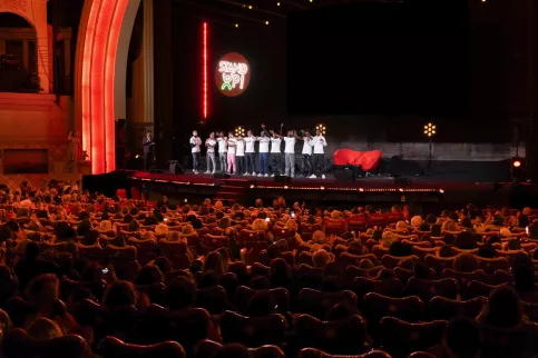 Vue de la scène et du public depuis le fond de la salle. Sur scène, un groupe de jeunes en tee-shirts blancs